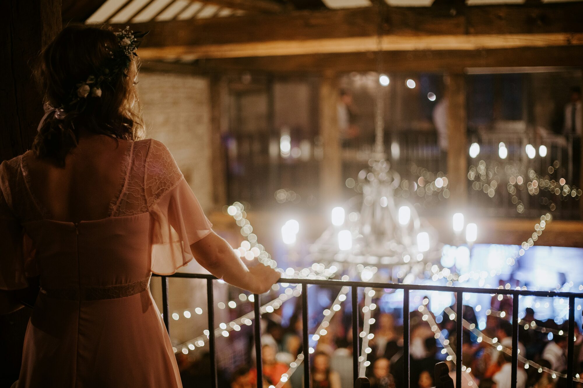 A bride pausing on a balcony, looking out over her reception as guests celebrate below.