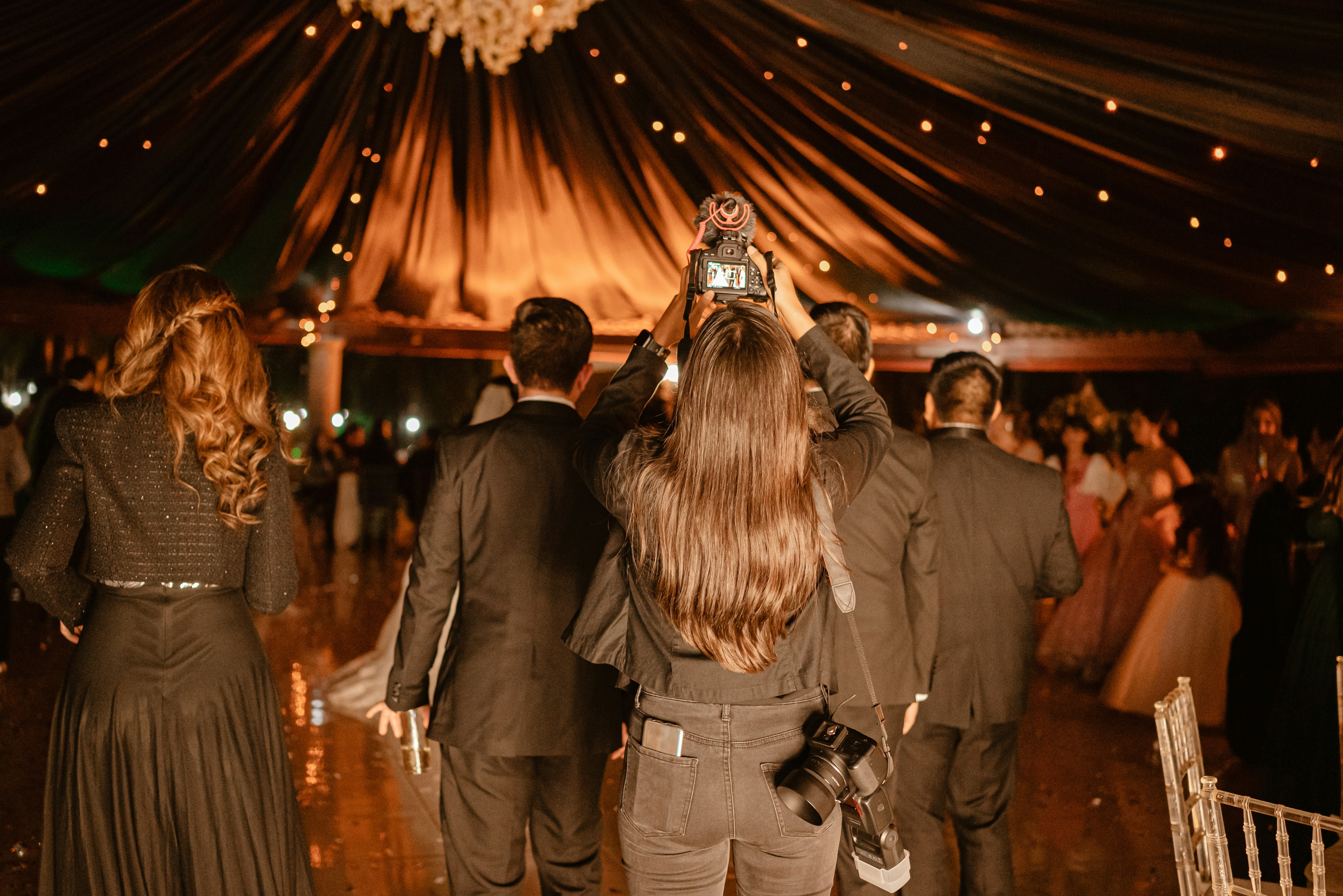 A wedding photographer captures a moment under a draped tent canopy strung with chandeliers and warm string lights.